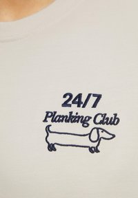 Lichtgrijze katoenen T-shirt met marineblauwe geborduurde tekst: "24/7 Planking Club" en een illustratie van een teckel. Gladde textuur.