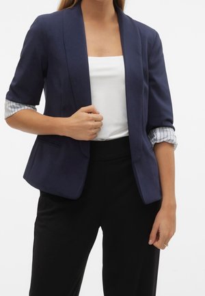 Femme portant un blazer bleu marine avec des manches rayées retroussées, un haut blanc et un pantalon noir, tenant le revers du blazer d'une main.