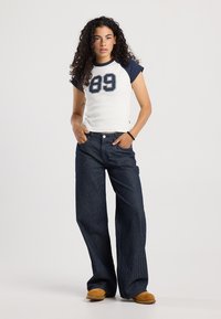 Camisa blanca de manga corta con detalles en azul marino y un gran estampado de "89", combinada con pantalones de mezclilla de pierna ancha en azul oscuro con rayas verticales y cierre de botón.