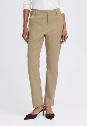 Beige maßgeschneiderte Hosen mit geradem Bein, Vordertaschen und dezenten Nähdetails, kombiniert mit dunkelbraunen spitzen Absätzen.