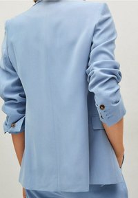 Svetle modrý na míru šitý blazer s ohrnutými rukávy, který má hnědé knoflíkové detaily a hladkou texturu látky. Zadní pohled ukazuje čisté linie.