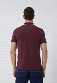 Burgunderfarbendes Poloshirt mit kurzen Ärmeln, weißem Kragen und Akzenten an den Ärmeln, aus glattem Stoff, getragen mit dunkelblauen Jeans. Einfarbiger Hintergrund.