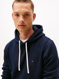 Σκούρο μπλε hoodie φτιαγμένο από μαλακό ύφασμα, με μπροστινή τσέπη, λευκές κορδόνες και ένα μικρό λογότυπο στο στήθος. Απλόν φόντο.