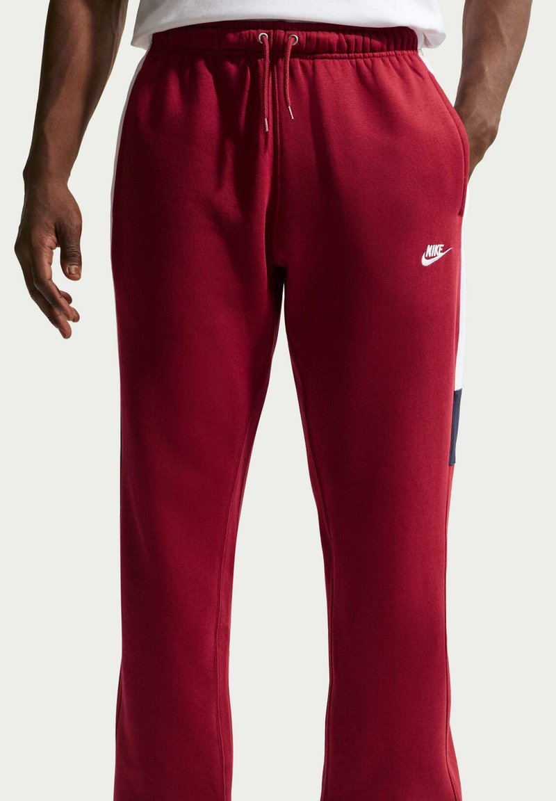 Uomo che indossa pantaloni della tuta Nike rossi con logo bianco e vita elastica con coulisse, mani rilassate lungo i fianchi.