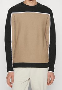 Maglione lavorato a maglia con un design a blocchi di colore, corpo beige, maniche nere e dettagli bianchi lungo le cuciture. Collo rotondo, tessuto testurizzato.
