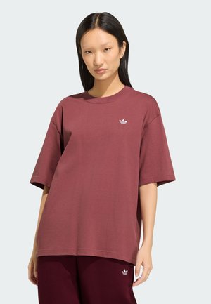 Ung kvinde med glat sort hår iført en oversize vinrød Adidas T-shirt og matchende bukser, stående mod en ensfarvet lys baggrund.