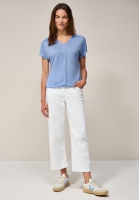 Un haut bleu clair à manches courtes et col en V, associé à un pantalon large blanc et des baskets beiges avec des accents bleus. Design simple, texture douce.