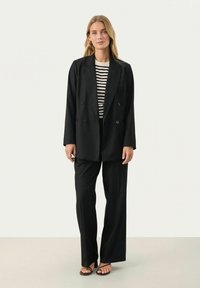 Černý dvouřadý blazer na černém a bílém pruhovaném tričku, doplněný černými širokými kalhotami a černými sandály, na neutrálním pozadí.