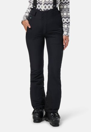 Femme portant un pantalon de ski isolé noir avec des bretelles réglables et des bottes de ski noires sur fond blanc.