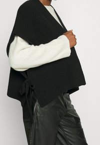 Czarny dzianinowy narzutka z krótkimi rękawami, z ozdobnym zapięciem po boku. Noszona na białym swetrze z prążkami i czarnych skórzanych spodniach.