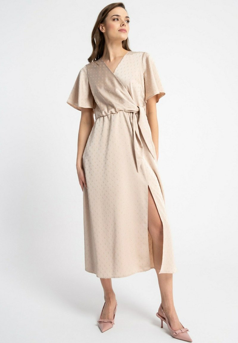 Beżowa sukienka w stylu kopertowym z krótkimi falbankowymi rękawami, fakturowana, z rozcięciem po boku i dopasowanymi różowymi szpilkami. Miękki, lekki materiał.