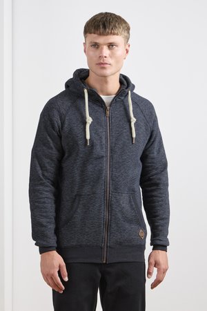 Sweat à zip couleur bleu marine foncé avec une texture douce, cordons de serrage crème, poches latérales et un petit logo sur le bas à gauche. Coupe décontractée.