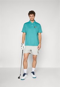 Türkiz színű pólóing, gallérral, rövid ujjú, hímzett részletekkel. Szürke rövidnadrággal és fehér golfcipővel párosítva, navy kiegészítőkkel.