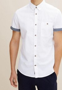 Camisa de botones blanca de manga corta con acentos de denim en los puños. Cuenta con un bolsillo delantero y botones marrón oscuro. Textura suave.