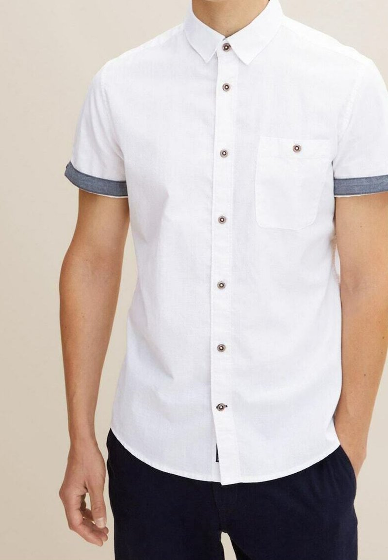 Camisa de botones blanca de manga corta con acentos de denim en los puños. Cuenta con un bolsillo delantero y botones marrón oscuro. Textura suave.
