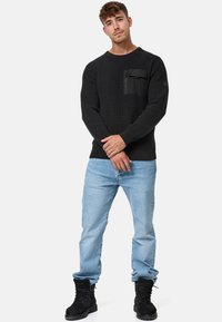 Pull en texture noire avec une poche poitrine zippée, associé à un jean bleu clair et des bottes noires à lacets. Le mannequin se tient dans une pose neutre.
