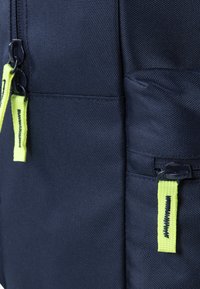 Sac à dos navy avec un tissu texturé, doté de zippers noirs et de tirettes jaunes vives. Comprend deux poches latérales zippées.