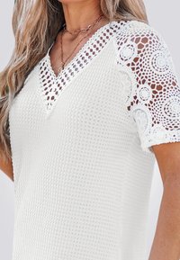 Bílý texturovaný top s výstřihem do V, který má krajkové háčkované rukávy a složité vzory podél okrajů. Prodyšný design.