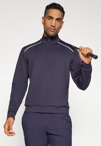 Pull-over bleu marine à zip quart en tissu doux et extensible, avec des rayures blanches sur les épaules, associé à un pantalon assorti ; mannequin tenant un club de golf.