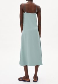 Langes, ärmelloses Kleid aus einem gedämpften grünen Stoff, mit geradem Schnitt und dünnen Trägern. Tragen mit marineblauen Flip-Flops. Glatte Textur.