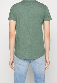 Osoba w krótkim zielonym podkoszulku i jasnoniebieskich dżinsach, pokazana z tyłu na jednolitym tle.