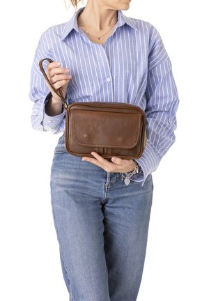 Donna con camicia a righe blu e jeans che tiene una borsa da polso in pelle marrone con una tasca frontale e un cinturino da polso.