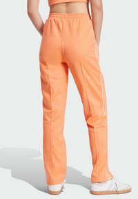 Orange athletic pants with a relaxed fit, featuring white side stripes, an elastic waistband, and smooth fabric. Worn with white sneakers.