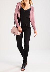 Cárdigan rosa claro sobre una blusa negra, combinado con jeans skinny negros. El conjunto se complementa con un pequeño bolso cruzado rosa y sandalias negras de tacón.