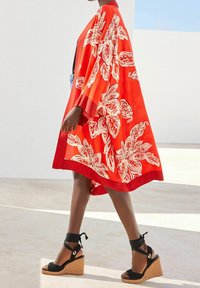 Kimono rouge avec motifs floraux blancs, tissu semi-transparent et larges manches. Porté avec des sandales à plateforme noires et des lanières autour de la cheville.