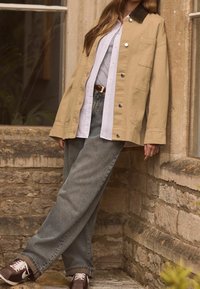 Beige puuvillane jakk pruunist kangast krae, valge nööbitav särk, hallid teksad ja tumedad spordijalatsid valgete detailidega. Seisab kivilise seina ääres.