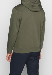 Hoodie i mörk olivgrön, tillverkad av mjukt tyg. Har en avslappnad passform, ribbad muddar och fåll. Synlig sömdetalj.