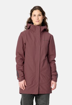Frau mit langen lockigen Haaren trägt eine knielange, bordeauxrote Regenjacke mit Kapuze über dunkler Hose vor schlichtem hellem Hintergrund.