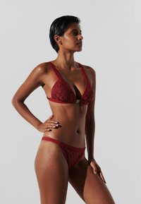 Raudonas bikinis, turintis trikampį viršų su subtiliu raštu, reguliuojamais diržais ir žemai nuleidžiamais kelniukais su atitinkamu dizainu.