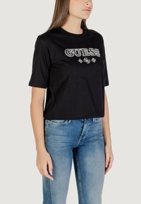T-shirt cropped nera in cotone, con un audace logo "GUESS" in bianco. Maniche corte, collo rotondo e vestibilità comoda. Abbinata a jeans blu.