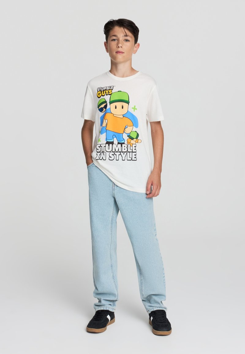 T-shirt bianco in cotone con un personaggio di cartone animato colorato, cappello verde e camicia arancione, abbinato a jeans azzurri chiari e sneakers nere.