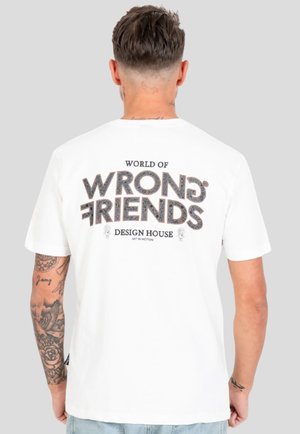 Mężczyzna z tatuażami w białej koszulce z napisem "World of Wrong Friends Design House" na plecach na jednolitym szarym tle.