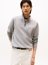 Sweat à zip gris avec col montant, porté sur une chemise à rayures. Associé à un jean blanc et une ceinture tissée noire. Design simple.