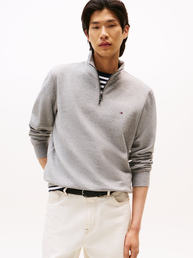 Sweat à zip gris avec col montant, porté sur une chemise à rayures. Associé à un jean blanc et une ceinture tissée noire. Design simple.