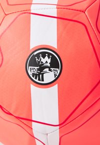 Ballon de football rouge avec une surface texturée, présentant une bande blanche et un logo circulaire avec une couronne et une figure stylisée.