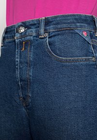 Jeans de mezclilla azul oscuro con costuras naranjas, que presentan un cierre de botón metálico y un pequeño detalle bordado de rosa en el bolsillo frontal.