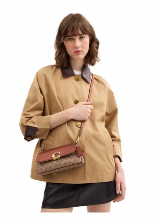 Mladá žena má na sebe béžovú bundu s tmavým golierom a manžetami, drží hnedú crossbody tašku a čiernu sukňu na bielom pozadí.