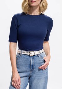 Laivastonsininen, ribbineulottu pusero lyhyillä hihoilla, yhdistetty korkeavyötäröisiin vaaleansinisiin farkkuihin ja valkoiseen punottuun vyöhön, jossa on pyöreä solki.