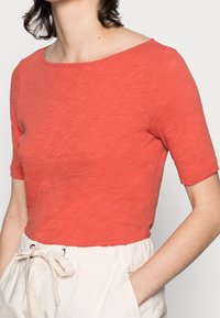 Top corallo a maniche corte realizzato in materiale morbido e textured con scollatura a barca. Abbinato a pantaloni color crema con elastico in vita e coulisse.