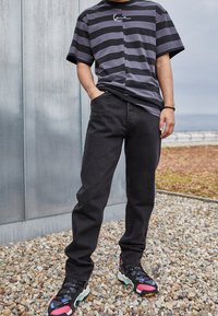Henkilö, jolla on päällään musta-harmaa raidoitettu t-paita, mustat farkut ja värikkäät kengät, seisoo soratiellä metalliseinän vieressä ulkona.