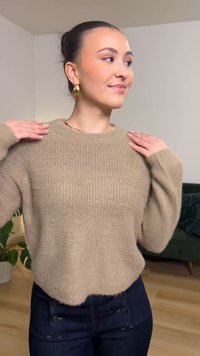 Beiger Strickpullover mit geripptem Muster, Rundhalsausschnitt und überschnittenen Schultern, kombiniert mit dunklen Jeans mit Knopfakzenten.