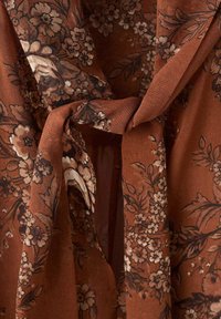 Brązowa tkanina w kwiaty o fakturowanej powierzchni, z misternymi czarnymi i kremowymi kwiatami. Tkanina związana w supeł, tworząc płynny wzór.