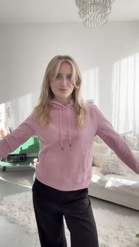 Noor blondidega naine, roosa kapuutsiga ja mustade pükstega, seisab avatud kätega heledas, moodsas elutoas.