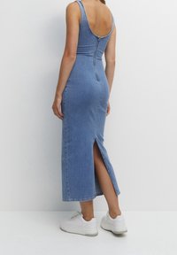 Robe maxi en denim bleu clair, sans manches avec un dos décolleté, dotée d'une fermeture éclair et d'une fente latérale. Associée à des baskets blanches.