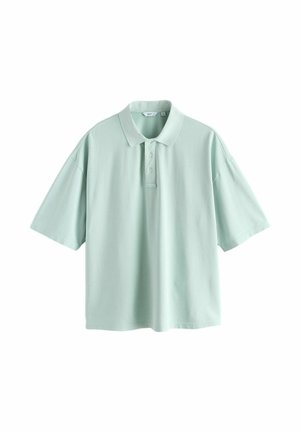 Fiatal férfi áll sima fal előtt, világoskék, túlméretezett pólót, porró rózsaszín rövidnadrágot és fekete, pántos szandált visel.