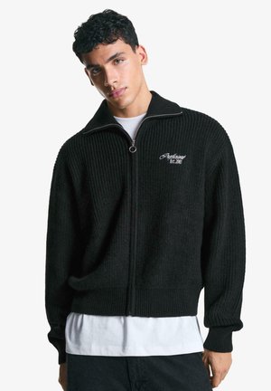 Black ribbed zip-up sweater with a high collar, featuring a white embroidered logo on the left chest and a textured material.